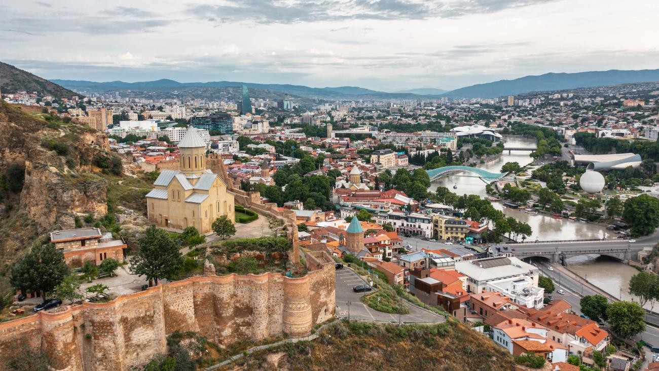 Panoramic view of Tbilisi from above showing Metekhi Church, the Peace Bridge, and the Kura River winding through the city