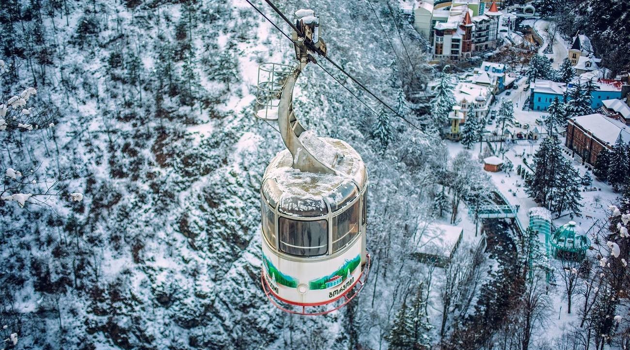 An aerial cable car gondola glides over a snow-blanketed forested valley and mountain town in Georgia