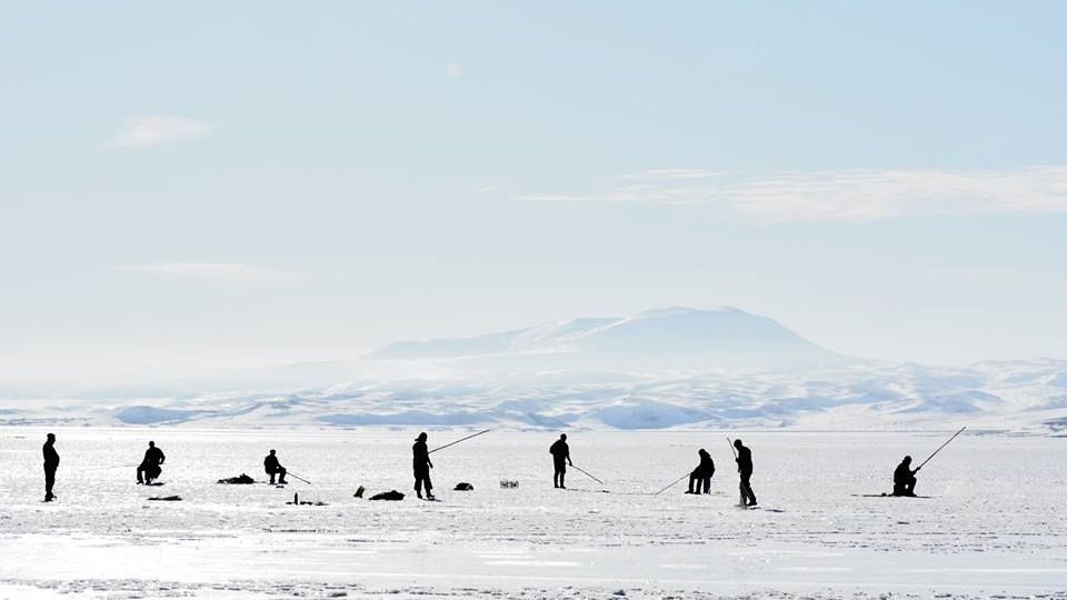 Silhouettes of ice fishermen with long rods standing on a vast frozen lake with snowy hills in the distance