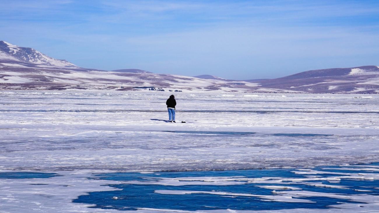 A lone person stands on a frozen lake with cracked blue ice stretching toward snow-dusted hills under a clear winter sky