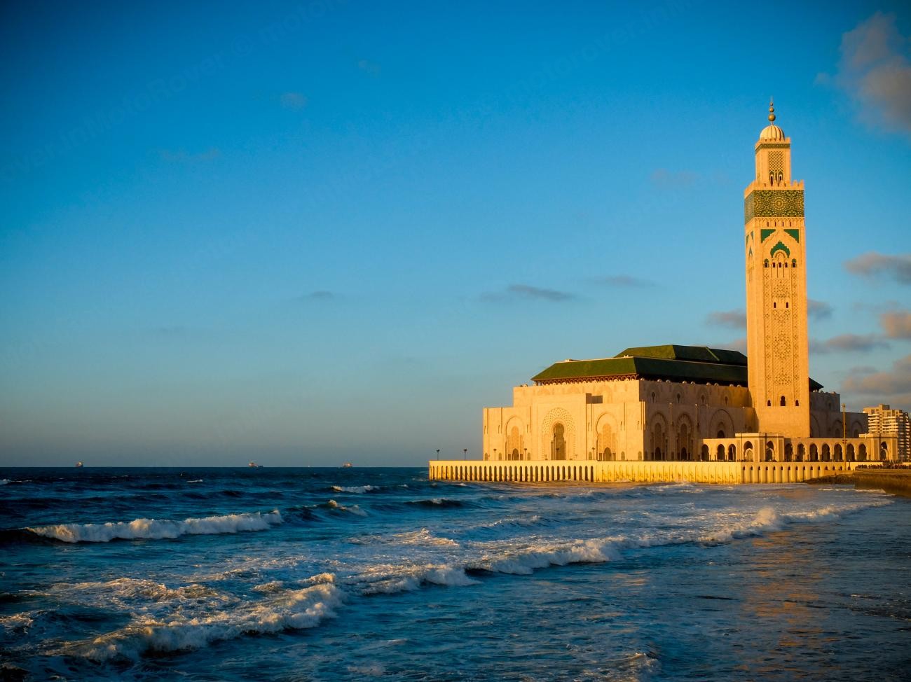 Hassan II Mosque in Casablanca glowing in golden light with Atlantic Ocean waves crashing at its base.