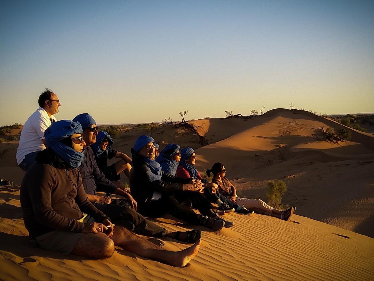 Group of travellers in blue headwraps sitting on Sahara sand dunes watching the sunset.