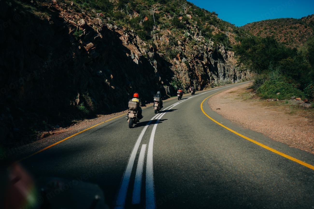 Motorcyclists riding through a dramatic rock-cut mountain pass with sweeping curves in the South African countryside.