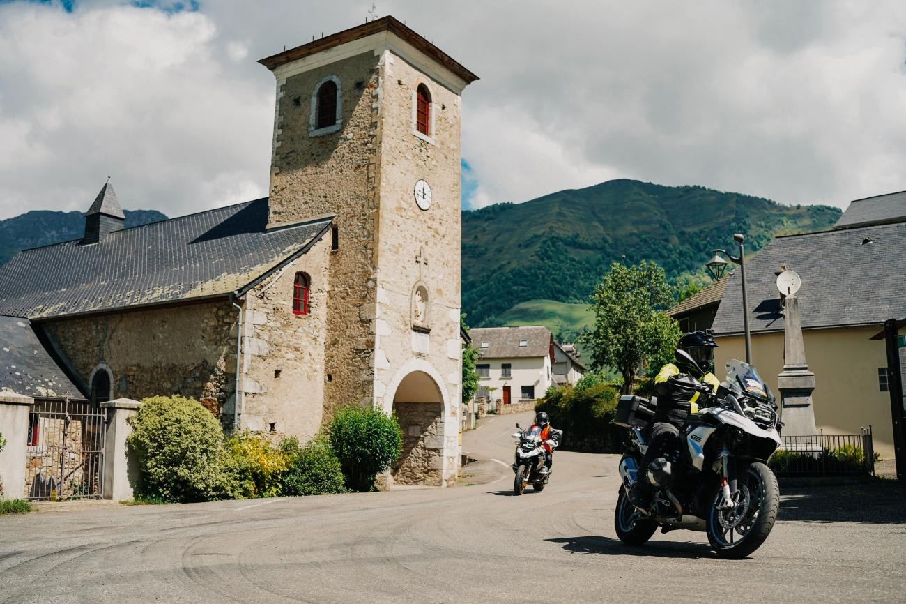 BMW GS adventure motorcycles parked beside a medieval stone church with a bell tower in a French Pyrenees village.