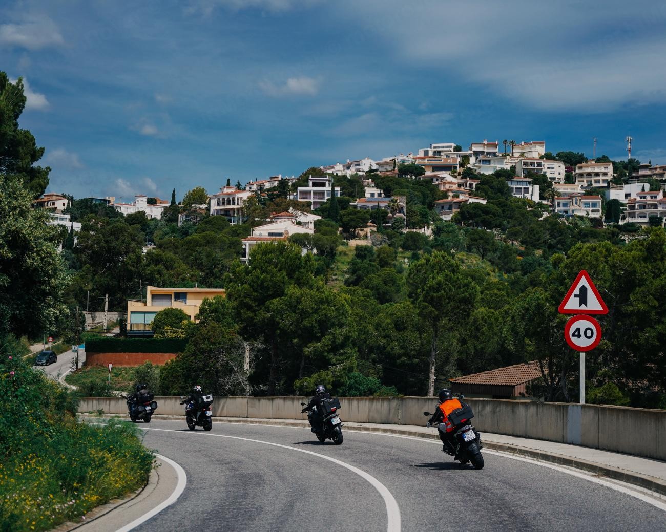 Group of motorcyclists navigating a curving road through a hillside Spanish coastal town.